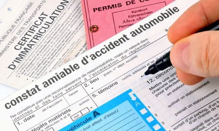 Main remplissant un constat amiable d’accident automobile avec des documents administratifs en arrière-plan